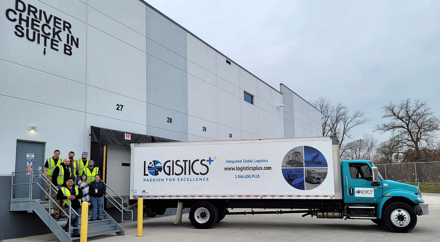 A group of Logistics Plus employees wearing reflective yellow vests stands together in front of a large Logistics Plus trailer at the loading dock.